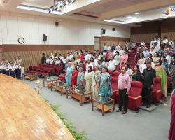 Large group of students and parents standing in a school auditorium during a ceremony, with a wooden stage on the left.