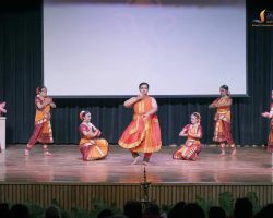 Group of young Bharatanatyam dancers in orange-red costumes performing on stage, with a central dancer mid-move and others posed around her.
