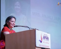 Smiling woman in a red outfit speaking at a podium with a microphone, Jaipuria School Lucknow banner on the podium and backdrop behind her.