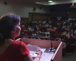 Speaker at a podium in a red top, reading notes into a microphone as a large audience sits in a dimly lit auditorium behind her.