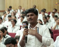 Student speaking into a handheld microphone in a crowded lecture hall of students wearing white shirts.