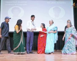 Group of people on a stage during an award ceremony, with a young man receiving a certificate from a woman in a red sari, others in colorful traditional outfits watching smilely.
