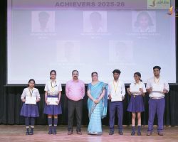 Group of students in uniform on a stage, receiving certificates at an awards ceremony, with 'ACHIEVERS 2025-26' banner in the background.