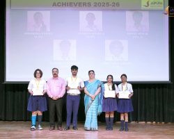 Group of students and teachers on a stage posing with certificates at an awards ceremony, with a projected backdrop reading 'Achievers 2025-26'.