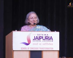 Indian woman in a saree speaking at a podium with a 'Jaipuria School Lucknow' banner visible on the front panel