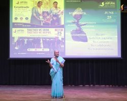 Woman in a light blue sari speaking on a stage with a large projected screen behind her showing posters and graphics.