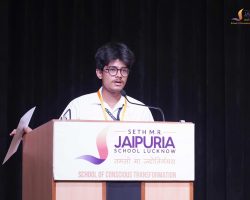 Young man speaker at a podium during a school event, wearing glasses and a badge, with a Jaipuria banner in the background.