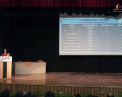 Speaker in a red saree at a podium on a stage, with a large projected slide showing a 'Comparison Table ICSE-2026' and an audience in front.