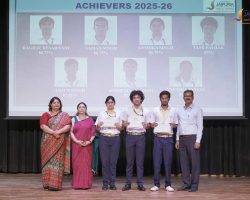 Group of students in uniform holding certificates on stage during an award ceremony, with teachers beside them and a projected backdrop reading 'Achievers 2025-26'.