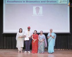 Six people on a stage pose with a certificate and medals after an award ceremony, backdrop shows 'Excellence in Dramatics and Oration'.