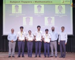 Group of students in uniform on stage receiving certificates at an awards ceremony, with teachers beside them and a banner reading 'Subject Toppers: Mathematics' in the background.