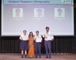 Group of three female students and one male student, plus a teacher, posing on stage with certificates after a geography toppers ceremony.