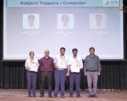 Five people stand on a stage during an award ceremony; three students in the center hold certificates and wear medals, flanked by two adults, with a presentation slide behind reading 'Subject Toppers: Computer'.