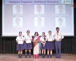 Group of students on a stage posing with certificates and medals during a school awards event; a projector screen behind shows 'Subject Toppers: Political Science'.