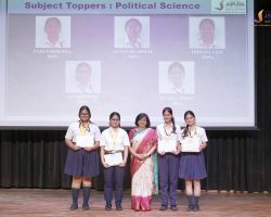 Group of female students on stage receiving certificates at a school award ceremony with a large projected screen in the background showing student toppers names.