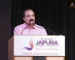 Man in a pink shirt giving a speech at a podium with microphones, Jaipuria School Lucknow banner visible on the front.