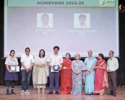 Group of students and staff posing with awards on a school stage during an achievers ceremony, with a projector screen in the background.