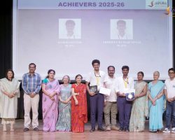 Group of students and teachers on stage during an award ceremony, holding plaques and certificates with a projection screen reading 'Achievers 2025-26' in the background.