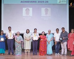 Group of adults on stage posing with trophies and certificates at an awards ceremony; screen behind reads 'ACHIEVERS 2025-26' with two portrait photos above them.