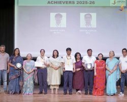 Group of award recipients on stage holding plaques and certificates, with a screen reading 'ACHIEVERS 2025-26' behind them.