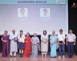 Group of students and adults posed on a school stage after an awards ceremony; a student holds a trophy and another holds a certificate, with a backdrop reading 'Achievers 2025-26'.