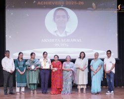 Group of people on a stage posing with awards during a ceremony, a large screen behind shows a portrait and the name Akshita Agrawal with 99.75%.