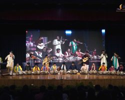 Wide view of a school concert: students on stage playing tabla, violin, sitar, keyboard and guitar with a large backdrop screen and seated audience in front.