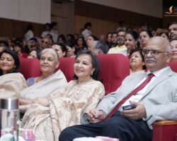 Front row of attendees at a formal event: an elderly woman in a beige sari and an older man in a gray suit with a red tie, smiling in red auditorium seats, with a crowded audience in the background.