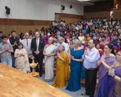 Diverse group of people standing with folded hands during a ceremony in a large auditorium, with a lit brass lamp in the foreground.
