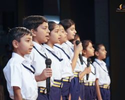 Group of schoolchildren in white shirts sing into handheld microphones on a stage, lined up in a row.