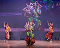 Group of young female dancers in vibrant traditional costumes performing a classical Indian dance on a stage with a wooden floor and a dark backdrop.