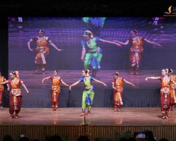 Group of dancers in bright traditional Indian outfits performing on stage in front of a large LED screen backdrop.