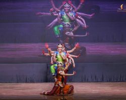 Colorful Indian classical dance on stage with a front dancer kneeling and others stacked behind her in a coordinated pose.