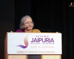 Elderly woman at a podium delivering a speech, wearing a purple sari, with a microphone in front of her and a banner reading 'JAIPURIA School Lucknow' in the foreground.