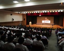 Speaker at a podium on stage addressing a crowded school assembly in a large auditorium filled with students in white uniforms.