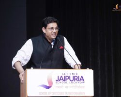 Man in glasses delivering a speech at a podium on a stage, with a Jaipuria School Lucknow banner on the lectern behind him.
