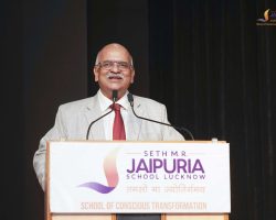 Man in a light gray suit and red tie speaks at a podium with two microphones; backdrop shows a banner for Jaipuria School Lucknow.
