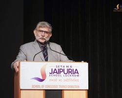 Older man in a gray suit delivers a speech at a podium with a Jaipuria School banner in front of a dark backdrop.