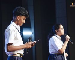Two students in white shirts on stage, each holding a microphone and reading from notes as they present.
