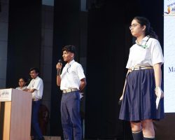 Students in school uniforms speaking on stage, one holding a microphone near a podium with a banner in the background (Jaipuria School Lucknow).
