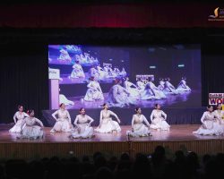 Group of female dancers in cream dresses perform a synchronized stage dance on a theatre stage, with a large LED screen showing the same performance behind them.