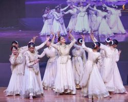 Group of women in cream dresses with gold trim performing a synchronized dance on stage, with a large screen displaying them in the background.