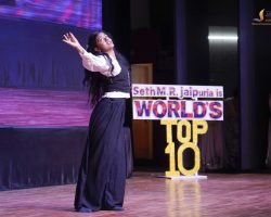 Young woman performing on a dimly lit stage with arms raised, wearing a white blouse and black skirt, beside a sign reading 'WORLD'S TOP 10'.