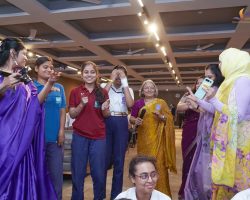 Group of students and teachers smiling and conversing at a school event, some in purple saris, indoors under ceiling lights.