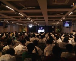 Large audience of students in white shirts seated in a hall, facing a stage with a speaker at a podium and screens on either side displaying the presenter.