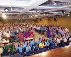 Large group of students in colorful uniforms cheering with hands raised in a school auditorium during an event.