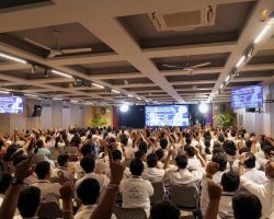 Large conference hall filled with people seated and raising hands, facing a stage with screens displaying a presentation titled 'Community Collaboration'.