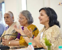 Panel of diverse adults at a conference, hands raised in prayer, microphones on the table in front of them (Jaipuriya logo top-right).