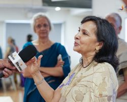 Woman in a beige patterned outfit being interviewed, speaking into a microphone with reporters in the background.