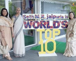 Four Indian women in beige sarees pose beside a colorful banner that reads "Seth M.R. Jaipuria is WORLD'S TOP" outside a building with glass doors and plants nearby.